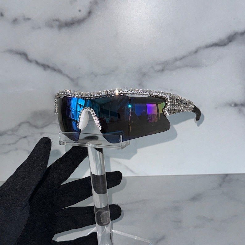 Pu&ograve; includere: Un paio di occhiali da sole con montatura argentata e lenti blu. La montatura &egrave; decorata con strass.