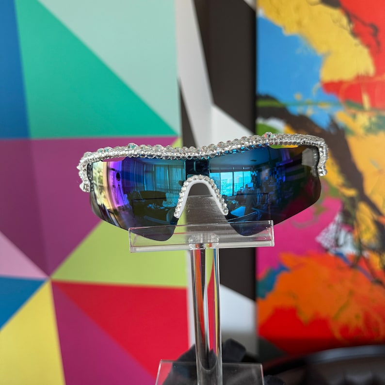 Pu&ograve; includere: Un paio di occhiali da sole blu a specchio con montatura bianca decorata con strass.