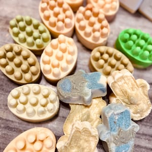 May include: A collection of handmade soap bars in various colours and shapes. The bars are made with natural ingredients and have a variety of textures, including smooth, bumpy, and textured. Some of the bars are shaped like hands, while others are rectangular.
