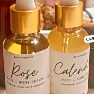 May include: Two glass bottles with gold lids and labels. The bottles contain a clear liquid. The labels read "Rose Face + Body Serum" and "Calendula Face + Body Serum". Both bottles are 30 ml or 1 oz.