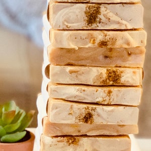 Puede incluir: Una pila de 12 barras de jabón hechas a mano con un aspecto marmoleado blanco y marrón claro. Las barras de jabón tienen un brillo sutil y están decoradas con pequeñas motas marrones.