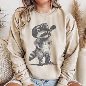 May include: A beige sweatshirt with a graphic print of a raccoon wearing a cowboy hat and boots. The raccoon is standing with its arms crossed and has a serious expression on its face.