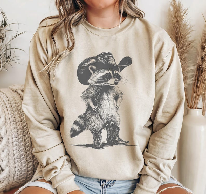 May include: A beige sweatshirt with a graphic print of a raccoon wearing a cowboy hat and boots. The raccoon is standing with its arms crossed and has a serious expression on its face.
