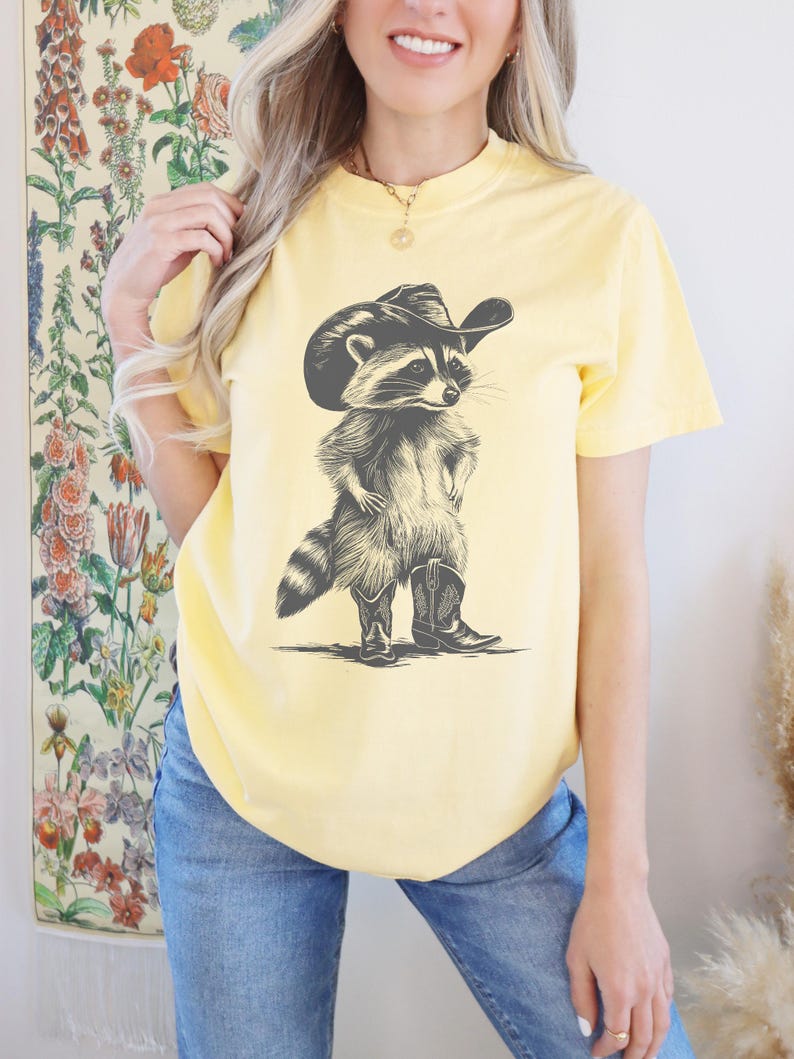 Pu&ograve; includere: Una maglietta gialla con una grafica di un procione che indossa un cappello da cowboy e stivali.