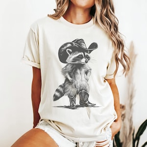 Pu&ograve; includere: Una donna indossa una maglietta color crema con un'illustrazione in bianco e nero di un procione che indossa un cappello da cowboy e stivali.
