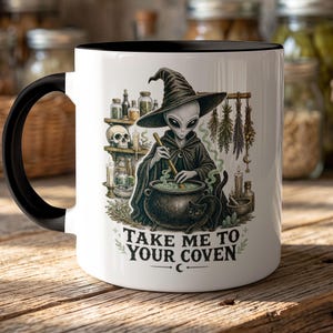 May include: A white ceramic mug with a black handle and rim featuring a graphic of an alien wearing a witch hat and cloak, stirring a cauldron with a black cat nearby. The design includes shelves with bottles, a skull, and the text TAKE ME TO YOUR COVEN.