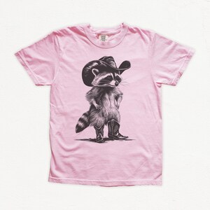 Pu&ograve; includere: Una maglietta rosa con un'illustrazione in bianco e nero di un procione che indossa un cappello da cowboy e stivali.