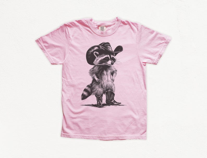 Pu&ograve; includere: Una maglietta rosa con un'illustrazione in bianco e nero di un procione che indossa un cappello da cowboy e stivali.