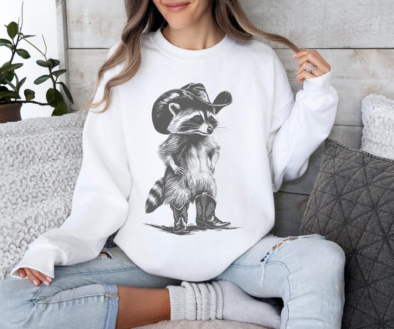 May include: White sweatshirt with a black and white illustration of a raccoon wearing a cowboy hat and boots.