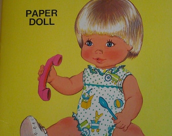 May include: A paper doll book featuring a baby doll with blonde hair and blue eyes. The doll is wearing a blue and white polka dot dress and is holding a pink phone. The book cover is yellow with the title "Baby this 'n that" and the text "Paper Doll".