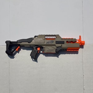 May include: A tan and black Nerf toy blaster featuring bright orange accents on the barrel, trigger, and stock. The plastic toy rifle has a tactical handle on top, a textured grip, and the Nerf logo clearly embossed on the side of the main body.