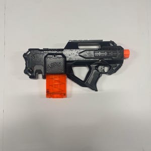 May include: A black and gray Nerf toy gun with an orange tip and an orange magazine. The toy gun has a tactical design with a handle and a sight. The Nerf logo is visible on the side of the gun.