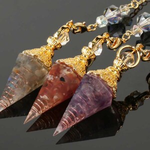May include: Three gemstone pendulums with gold-colored metal accents and chains. The pendulums are in shades of gray, red, and purple. Each pendulum has a pointed tip and is wrapped with copper wire.