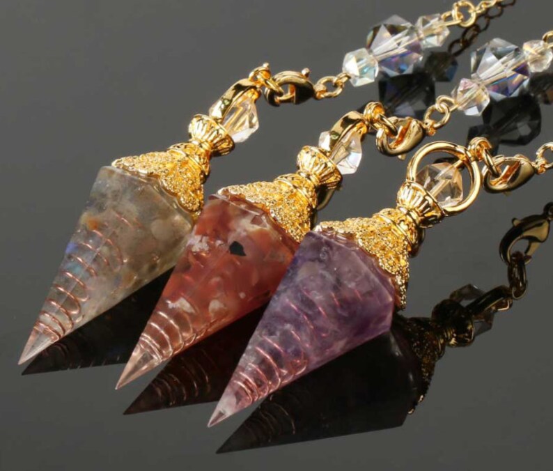 May include: Three gemstone pendulums with gold-colored metal accents and chains. The pendulums are in shades of gray, red, and purple. Each pendulum has a pointed tip and is wrapped with copper wire.