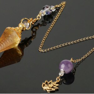 May include: A gold-colored pendulum with a faceted, amber-colored crystal point. The pendulum features a gold chain, a purple amethyst bead, and a decorative gold charm. Clear crystal beads adorn the top.