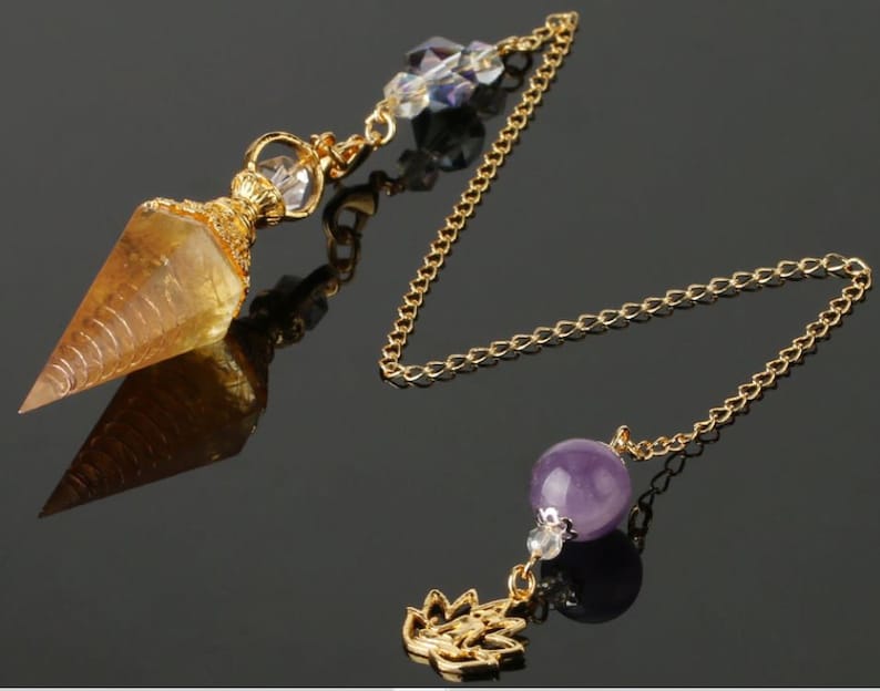 May include: A gold-colored pendulum with a faceted, amber-colored crystal point. The pendulum features a gold chain, a purple amethyst bead, and a decorative gold charm. Clear crystal beads adorn the top.