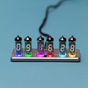 May include: A silver metal clock with seven clear glass tubes displaying the numbers 08:28. Each tube has a different colored light inside, including red, purple, green, blue, and yellow.
