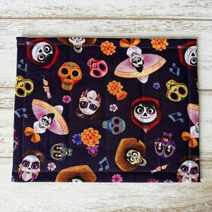 May include: A dark blue rectangular fabric with a repeating pattern of colorful sugar skulls, flowers, musical notes, and characters from the movie "Coco." The edges are neatly stitched, and the design is vibrant and detailed.