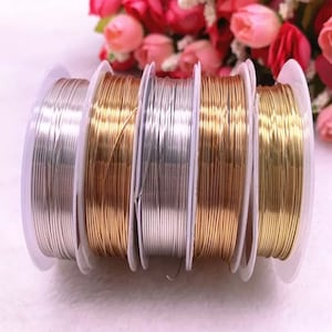 May include: Five spools of wire for jewellery making. The spools are arranged in a row, with the silver wire on the left, followed by gold, silver, gold, and silver. The spools are all the same size and have a white background.