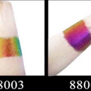 Op de afbeelding: Twee swatches van iriserende glitterpigment op de huid. De swatch links is regenboogkleurig, en de swatch rechts is paars. De swatches zijn gelabeld 88003 en 88004.