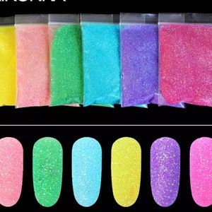 Puede incluir: Seis pequeñas bolsas de purpurina iridiscente en varios colores: amarillo, rosa, verde, azul, morado y rojo. La purpurina se muestra sobre una superficie negra.