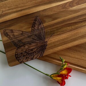 May include: A wooden chopping board featuring a laser-etched butterfly design. The butterfly is brown and black with intricate details. The board is made from natural wood and has a warm, rustic appearance.