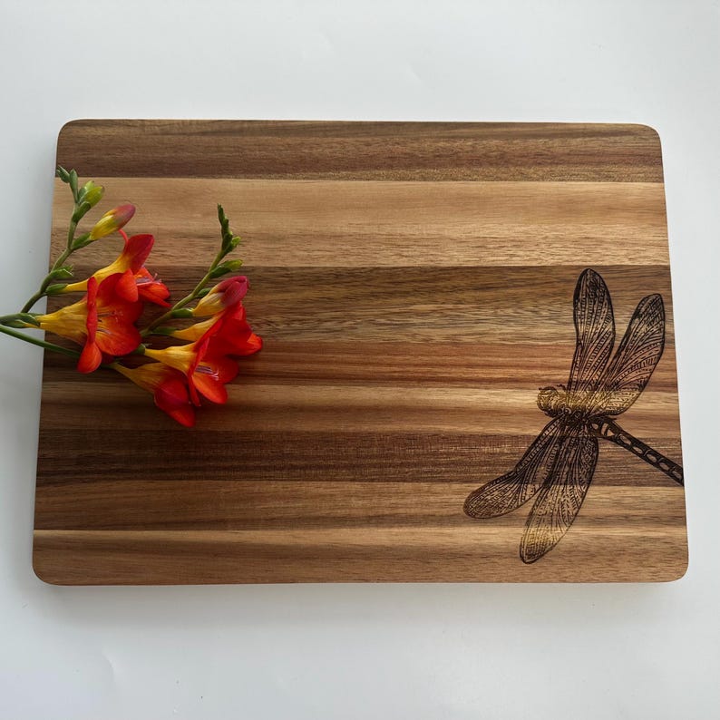May include: A wooden chopping board with a natural wood grain finish. The board features a dragonfly design engraved on the surface. The board is approximately 30 cm by 45 cm.