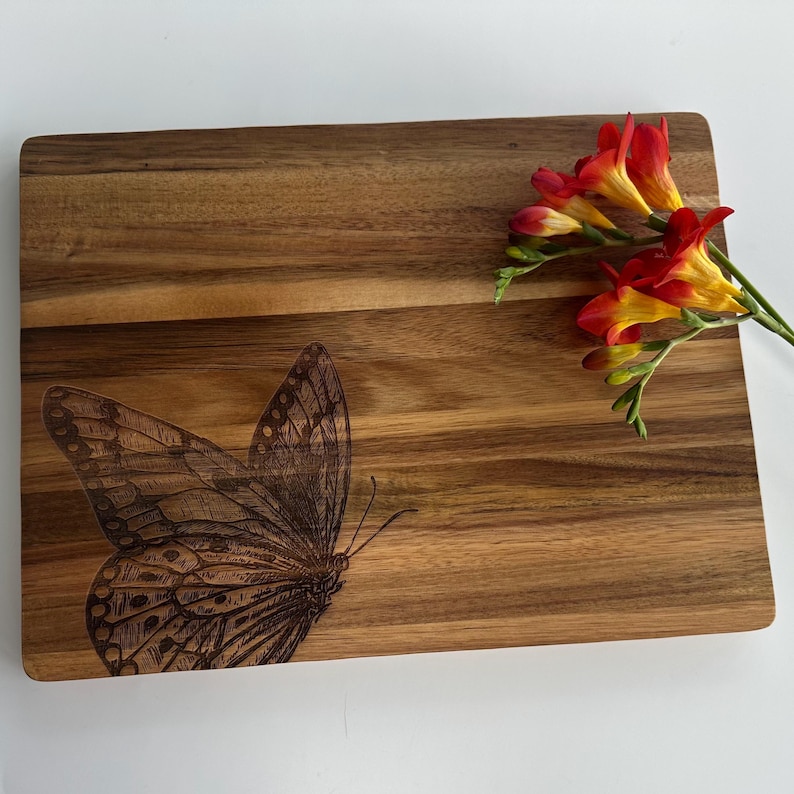 May include: A wooden chopping board with a laser-etched butterfly design. The board is brown and has a natural wood grain. There are red and yellow flowers on the board.