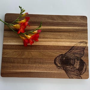 May include: A wooden chopping board with a natural wood grain finish. The board has a bee design engraved on the bottom right corner. A sprig of orange and yellow flowers is placed on the top left corner of the board.