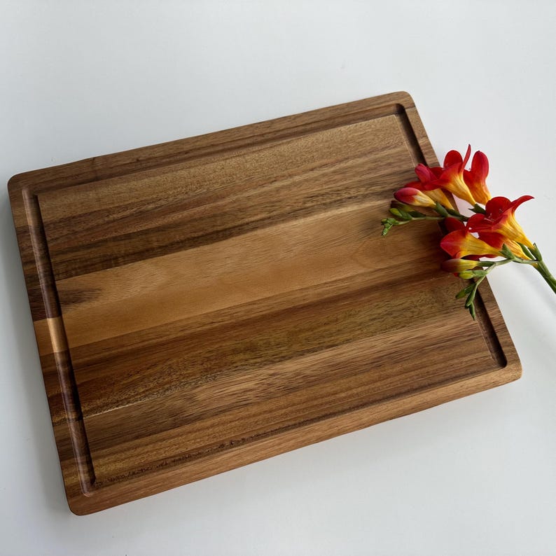 May include: A large, rectangular wooden chopping board with a natural wood grain finish. The board has a raised edge and a groove around the perimeter. A cluster of orange and yellow flowers is resting on the board.