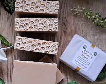 May include: An assortment of handmade soaps, some with a honeycomb pattern, others smooth. The soaps are light brown and beige, arranged on a wooden surface with green foliage. One soap is wrapped in white paper with text.