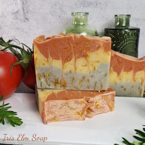 May include: Three bars of handmade soap with a layered design of orange, yellow, and gray hues. The soap is decorated with small, dried flower petals. The image also includes two red tomatoes and green glass bottles.