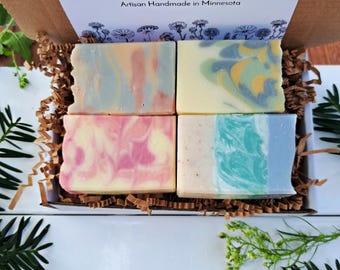 May include: A box set of four artisan soaps with swirled colors of pink, blue, yellow, and green. The soaps are nestled in brown crinkle paper inside a cardboard box. The box has a white label with the text "Iris Elm Soap" and "Artisan Handmade in Minnesota."