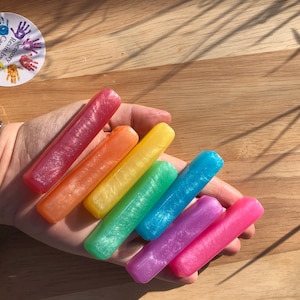 Peut inclure: Une main présente sept pièces de résine rectangulaires et colorées, disposées en arc-en-ciel. Les couleurs comprennent le rouge, l'orange, le jaune, le vert, le bleu, le violet et le rose. Les pièces ont un effet scintillant. Un autocollant indique "Sensory Resin by Carrie".