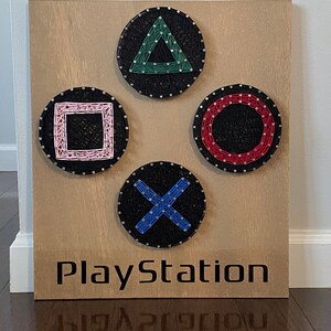 May include: A wooden board with four string art circles depicting the PlayStation controller symbols: a green triangle, a pink square, a red circle, and a blue X. The word "PlayStation" is written below the circles.