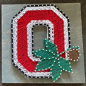 May include: A string art piece featuring a large letter "O" in red, white, and black string, with a green leaf and brown acorn design. The artwork is mounted on a gray wooden board, showcasing a handcrafted aesthetic.