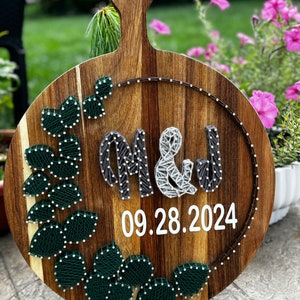 May include: A wooden round cutting board with a handle, decorated with string art. The string art features the letters M & J, a leafy design, and the date 09.28.2024.