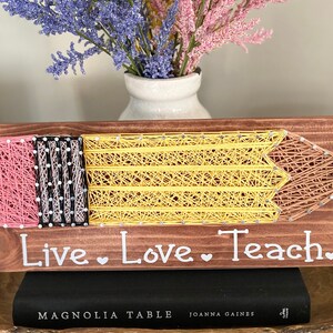 May include: Wooden pencil-shaped string art with the words "Live Love Teach" in white. The pencil features pink, black, yellow, and brown string. A vase of flowers sits above the art piece.