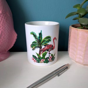May include: White ceramic mug with a colourful illustration of a flamingo, palm tree, and tropical flowers. The design features pink, green, and red hues. The mug is cylindrical and rests on a white surface.