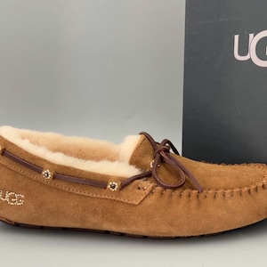 May include: Brown suede UGG slippers with a shearling lining and a decorative brown leather strap with gold accents. The slippers have a moccasin style toe and a UGG logo on the side.