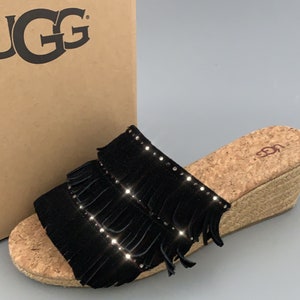 May include: Black suede wedge sandals with a cork platform and fringe detail. The sandals are adorned with rhinestones and have the brand name "UGG" on the cork platform.