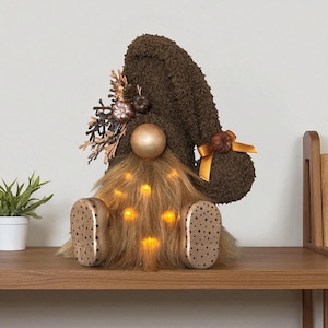 Peut inclure: Un gnome décoratif avec un chapeau marron texturé et une longue barbe brun clair. Le gnome a un nez doré et des chaussures dorées à pois marron. La barbe a de petites lumières aux tons chauds. Le chapeau est décoré de petites citrouilles et de feuilles décoratives.