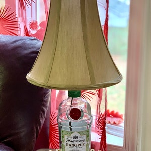 May include: A lamp made from a clear glass bottle with a green top and a red cap. The bottle is labeled "Tanqueray Rangpur Distilled Gin". The lamp has a beige fabric lampshade.