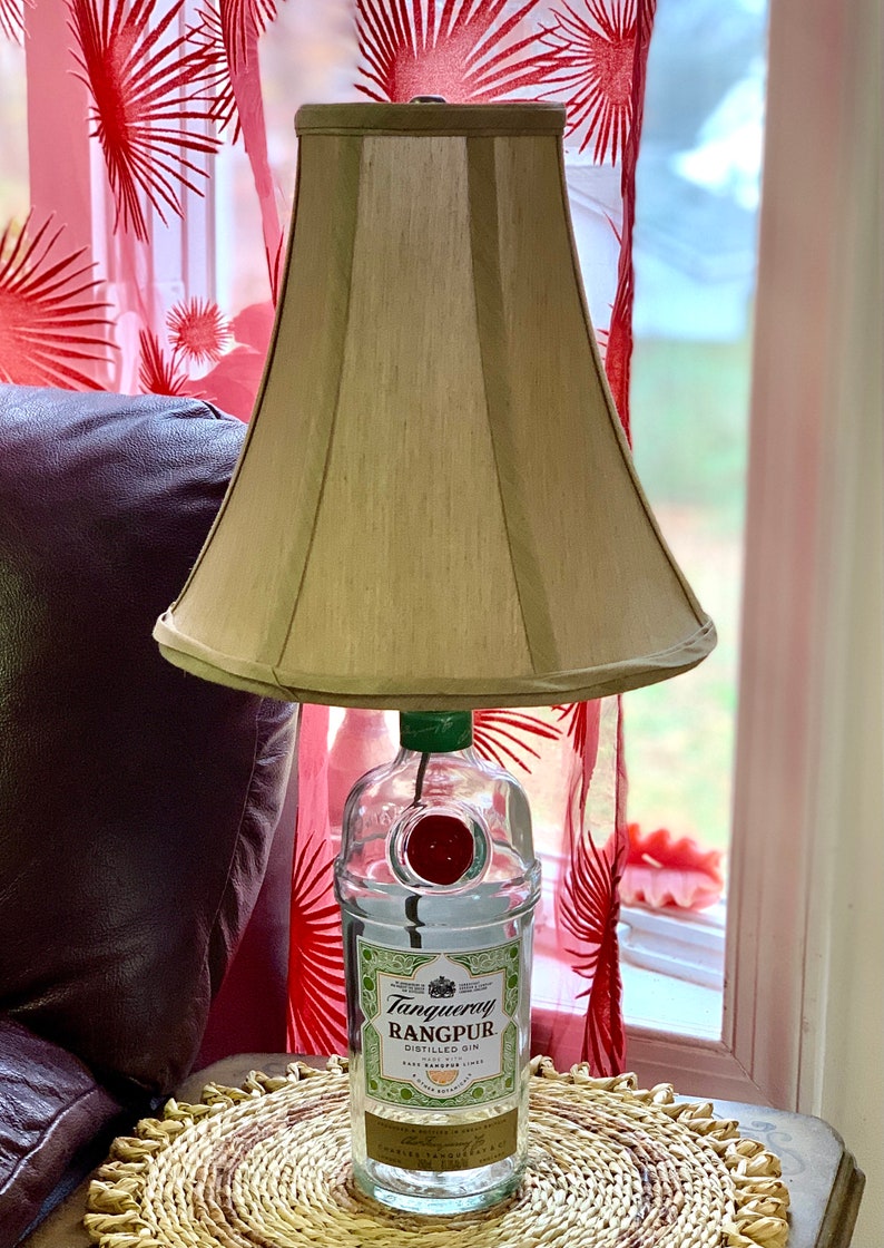May include: A lamp made from a clear glass bottle with a green top and a red cap. The bottle is labeled "Tanqueray Rangpur Distilled Gin". The lamp has a beige fabric lampshade.