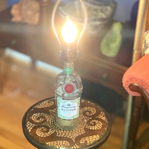 May include: A repurposed Tanqueray Rangpur gin bottle lamp with a clear glass shade and a light bulb turned on. The lamp is sitting on a small round table with a mosaic top.
