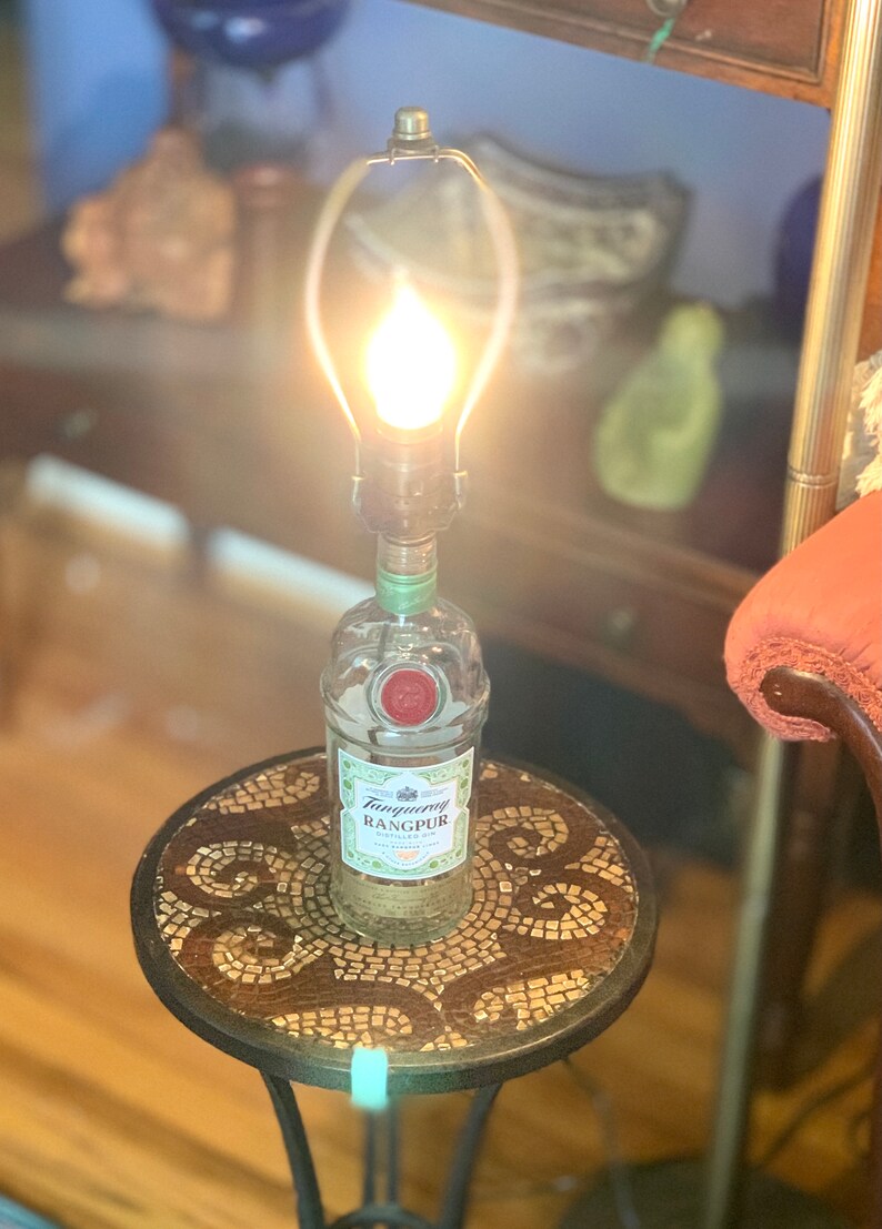 May include: A repurposed Tanqueray Rangpur gin bottle lamp with a clear glass shade and a light bulb turned on. The lamp is sitting on a small round table with a mosaic top.