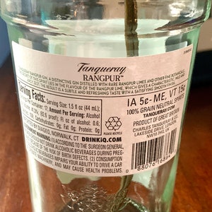 May include: A clear glass bottle of Tanqueray Rangpur gin with a white label. The label has the brand name, product description, serving facts, and a warning about drinking alcohol during pregnancy. The bottle is sitting on a wooden surface.