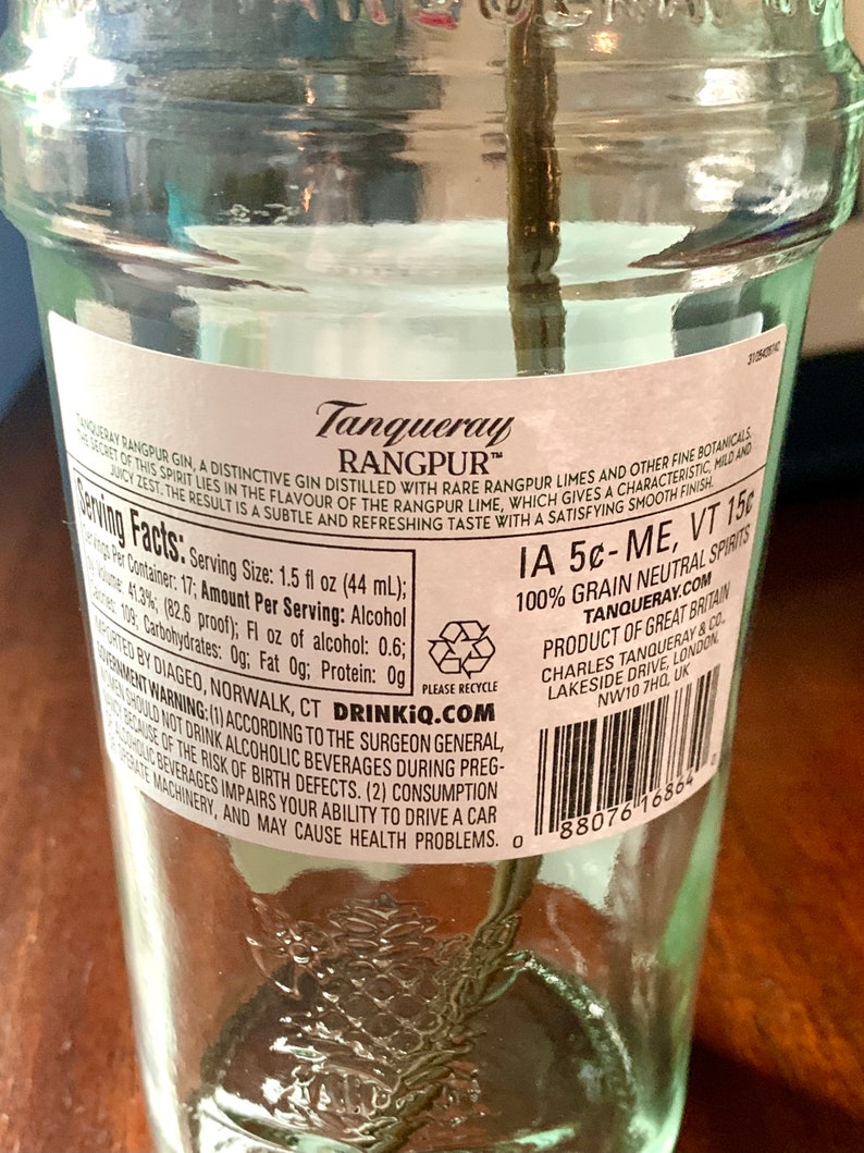 May include: A clear glass bottle of Tanqueray Rangpur gin with a white label. The label has the brand name, product description, serving facts, and a warning about drinking alcohol during pregnancy. The bottle is sitting on a wooden surface.