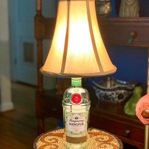May include: A repurposed Tanqueray Rangpur gin bottle lamp with a cream-colored lampshade. The bottle is clear glass with a green top and a red seal. The lamp is sitting on a small, round table with a mosaic tile top.