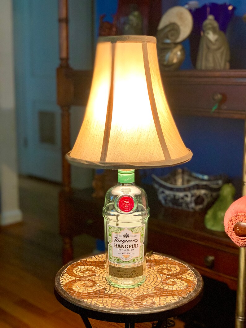 May include: A repurposed Tanqueray Rangpur gin bottle lamp with a cream-colored lampshade. The bottle is clear glass with a green top and a red seal. The lamp is sitting on a small, round table with a mosaic tile top.